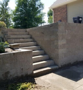a tall retaining wall with built-in stairs above a concrete patio or driveway and below a house with tall trees in the background