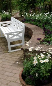 a white bench on a brick patio in front of a small retaining wall with white and dark purple flowers