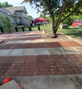 a large patio of light tan/gray concrete and red brick featuring landscaping including a tree, bushes, and a large decorative rock